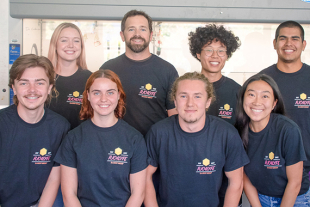 Members of the Costanzo Lab research team stand together in a laboratory wearing matching T-shirts while participating in a Cal Poly chemistry research project.