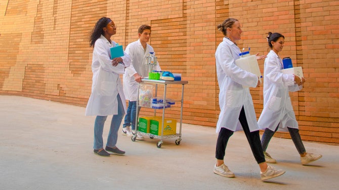 Researchers wearing white lab coats and carrying lab equipment walk past the Frost Center