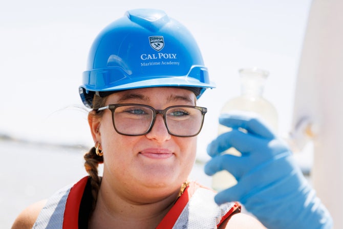 Oceanography student wearing a hard hat and life vest takes a water sample on a boat.